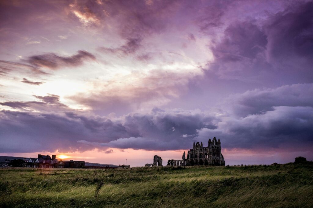 Silhouette des ruines gothiques de l'abbaye de Whitby s'élevant sur une colline herbeuse sous un ciel d'orage aux teintes violettes et pourpres lors du coucher du soleil.