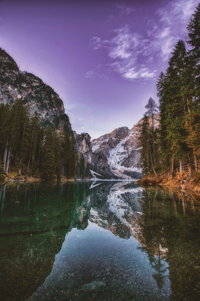 Paysage de montagne montrant un lac aux eaux cristallines reflétant parfaitement un sommet enneigé et une forêt de sapins sous un ciel aux teintes violettes.