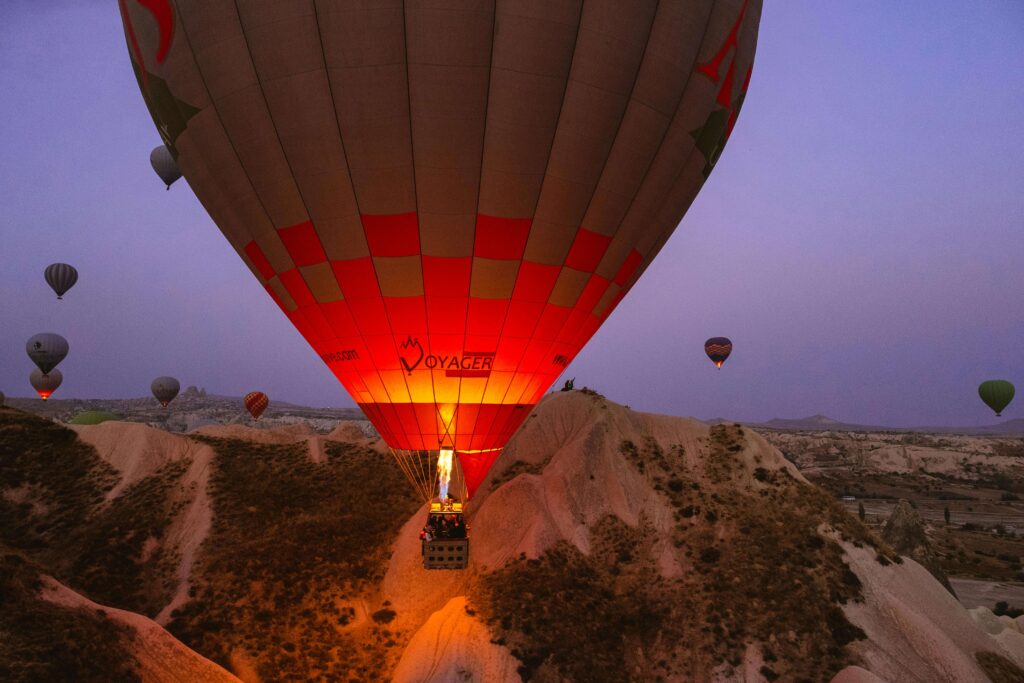 Gros plan sur la base d'une montgolfière illuminée par son brûleur au décollage, survolant les formations rocheuses de Cappadoce avec d'autres ballons au loin dans un ciel de fin de nuit.